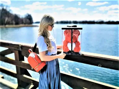 woman standing on a pier overlooking water, holding a luxury rose bear in gift box